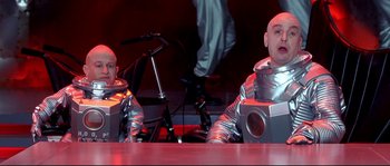 Movie still from “Austin Powers: The Spy Who Shagged Me” (1999), directed by Jay Roach – Two men in silver suits sitting in front of a microphone; Medium shot, Low angle
