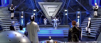 Movie still from “Austin Powers: The Spy Who Shagged Me” (1999), directed by Jay Roach – A group of people sitting on a stage in front of an audience; Wide shot, High angle