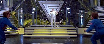Movie still from “Austin Powers: The Spy Who Shagged Me” (1999), directed by Jay Roach – A man standing in front of a stage with chairs and lights; Wide shot, Low angle