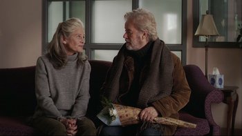 Movie still from “Away from Her” (2006), directed by Sarah Polley – An older man and woman sitting next to each other on a couch; Medium shot, Over the shoulder angle