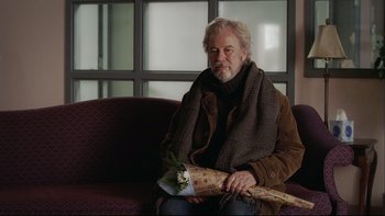 Movie still from “Away from Her” (2006), directed by Sarah Polley – An older man sitting on a couch holding a bouquet of flowers; Medium shot, Over the shoulder angle