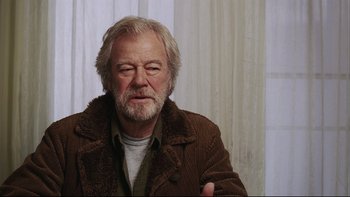 Movie still from “Away from Her” (2006), directed by Sarah Polley – An older man wearing a brown jacket is sitting in front of a curtain; Close Up shot, Over the shoulder angle