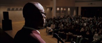 Movie still from “Stay” (2005), directed by Marc Forster – A man with glasses is speaking into a microphone in front of an audience; Close Up shot, Low angle