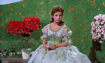 Movie still from “Babes in Toyland” (1961), directed by Jack Donohue – A woman in a dress sitting in front of a plant; Medium shot, Low angle
