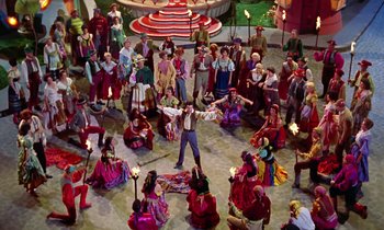 Movie still from “Babes in Toyland” (1961), directed by Jack Donohue – A group of people sitting in a circle and a person standing in front of them holding sparklers in their hands; Extreme Wide shot, High angle