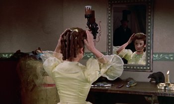 Movie still from “Babes in Toyland” (1961), directed by Jack Donohue – A woman in a yellow dress combing her hair in front of a mirror; Medium shot, Over the shoulder angle