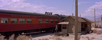 Movie still from “Bad Day at Black Rock” (1955), directed by John Sturges – A red and orange train traveling down train tracks next to a station; Extreme Wide shot, Low angle