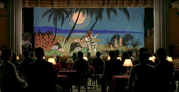 Movie still from “Bad Education” (2004), directed by Pedro Almodóvar – People are sitting in front of a mural of a man playing a guitar; Extreme Wide shot, Low angle
