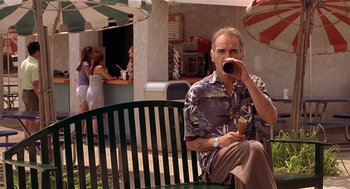 Movie still from “Bad Santa” (2003), directed by Terry Zwigoff – A man sitting on top of a wooden bench drinking a beverage; Medium shot, Over the shoulder angle