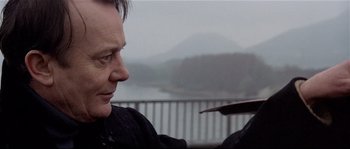 Movie still from “Bad Timing: A Sensual Obsession” (1980), directed by Nicolas Roeg – A man holding something in his hand while standing in front of a body of water; Close Up shot, Over the shoulder angle