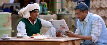 A man sitting at a table reading a newspaper to another man