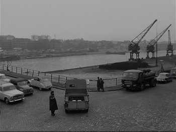 Movie still from “Band of Outsiders” (1964), directed by Jean-Luc Godard – A black and white photo of a man standing next to a car; Extreme Wide shot, High angle