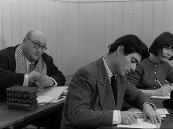Movie still from “Band of Outsiders” (1964), directed by Jean-Luc Godard – A man sitting at a table writing on a piece of paper next to another man; Medium shot, Over the shoulder angle