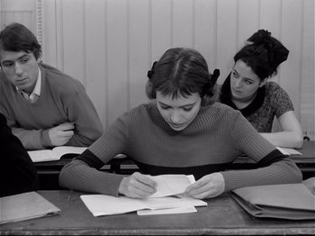 Movie still from “Band of Outsiders” (1964), directed by Jean-Luc Godard – A woman sitting at a table writing on a piece of paper; Medium shot, High angle