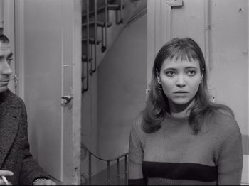 Movie still from “Band of Outsiders” (1964), directed by Jean-Luc Godard – A woman standing next to a staircase in a room; Medium shot, Over the shoulder angle