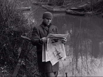 Movie still from “Band of Outsiders” (1964), directed by Jean-Luc Godard – A man standing by a body of water reading a newspaper; Medium shot, High angle