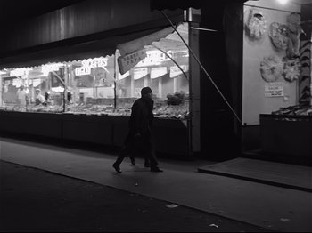 Movie still from “Band of Outsiders” (1964), directed by Jean-Luc Godard – A man walking down the street in front of a store; Wide shot, High angle