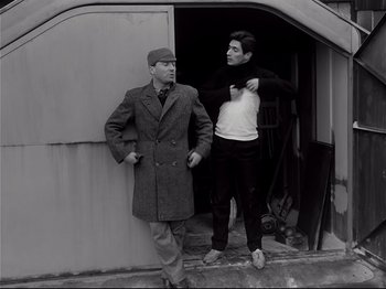 Movie still from “Band of Outsiders” (1964), directed by Jean-Luc Godard – Two men standing next to each other in front of a building; Medium shot, Low angle