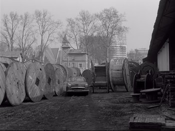 Movie still from “Band of Outsiders” (1964), directed by Jean-Luc Godard – An old car parked next to a bunch of wooden barrels; Extreme Wide shot, High angle