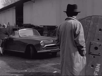 Movie still from “Band of Outsiders” (1964), directed by Jean-Luc Godard – A man in a trench coat and a hat standing in front of an old car; Wide shot, Over the shoulder angle