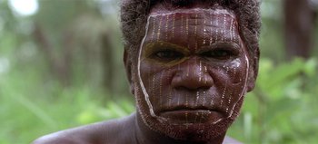 Movie still from “Baraka” (1992), directed by Ron Fricke – A man with a painted face is standing in the woods; Close Up shot, High angle