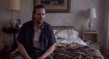 Movie still from “Barfly” (1987), directed by Barbet Schroeder – A man sitting on a bed in front of a painting; Close Up shot, Low angle