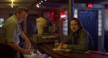 Movie still from “Barfly” (1987), directed by Barbet Schroeder – A man sitting at a table in front of an old suitcase; Medium shot, Over the shoulder angle