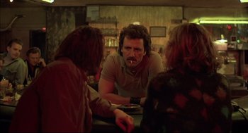 Movie still from “Barfly” (1987), directed by Barbet Schroeder – Three people sitting at a table in front of a man smoking a cigarette; Medium shot, Low angle