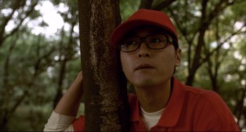 Movie still from “Barking Dogs Never Bite” (2000), directed by Bong Joon Ho – A man wearing a red hat and glasses leaning against a tree; Close Up shot, Low angle
