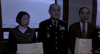 Movie still from “Barking Dogs Never Bite” (2000), directed by Bong Joon Ho – A man in uniform standing next to two other men; Medium shot, Over the shoulder angle