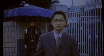 Movie still from “Barking Dogs Never Bite” (2000), directed by Bong Joon Ho – A man in a suit and tie standing in front of an officer; Medium shot, Low angle