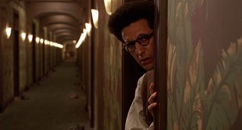 Movie still from “Barton Fink” (1991), directed by Joel Coen – A man with glasses leaning against a wall in a hallway; Medium shot, Over the shoulder angle