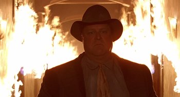 Movie still from “Barton Fink” (1991), directed by Joel Coen – A man in a hat and a suit standing in front of a fire; Medium shot, Low angle