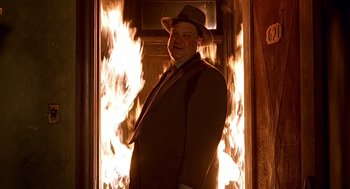 Movie still from “Barton Fink” (1991), directed by Joel Coen – A man in a suit and hat standing in front of a fire; Medium shot, Low angle