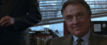 Movie still from “Basic Instinct” (1992), directed by Paul Verhoeven – An older man wearing a suit and tie in front of a bookshelf; Close Up shot, Over the shoulder angle