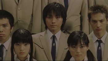 Movie still from “Battle Royale” (2000), directed by Kinji Fukasaku – A man in a suit and tie standing between two women in a suit; Close Up shot, Low angle