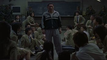 Movie still from “Battle Royale” (2000), directed by Kinji Fukasaku – A group of people sitting in front of a man in front of a chalkboard; Wide shot, High angle