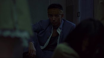 Movie still from “Battle Royale” (2000), directed by Kinji Fukasaku – A man with a bandage on his head sitting in a dark room; Medium shot, Low angle