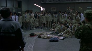 Movie still from “Battle Royale” (2000), directed by Kinji Fukasaku – A group of people in a room with a gun; Wide shot, High angle