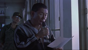 Movie still from “Battle Royale” (2000), directed by Kinji Fukasaku – A man holding a microphone while looking at a piece of paper; Close Up shot, Low angle