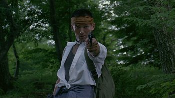 Movie still from “Battle Royale” (2000), directed by Kinji Fukasaku – A man with a bandana on his head points a gun at the camera; Medium shot, Low angle