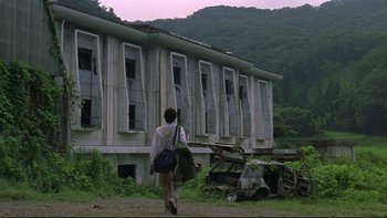 Movie still from “Battle Royale” (2000), directed by Kinji Fukasaku – A man walking down a dirt road past an abandoned building; Extreme Wide shot, Low angle