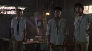 Movie still from “Battle Royale” (2000), directed by Kinji Fukasaku – A man standing in front of a table with papers on it; Medium shot, Low angle