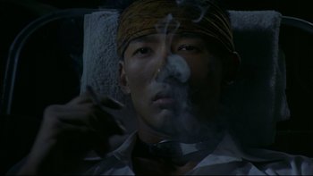 Movie still from “Battle Royale” (2000), directed by Kinji Fukasaku – A man smoking a cigarette while laying in a bed; Close Up shot, High angle