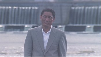 Movie still from “Battle Royale” (2000), directed by Kinji Fukasaku – An older man in a gray suit standing in front of a waterfall; Medium shot, Low angle