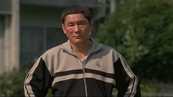 Movie still from “Battle Royale” (2000), directed by Kinji Fukasaku – A man in a black and white jacket standing in front of a building; Close Up shot, Low angle