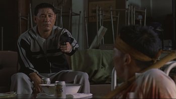 Movie still from “Battle Royale” (2000), directed by Kinji Fukasaku – A man sitting on a couch holding a remote control; Medium shot, Over the shoulder angle
