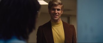 Movie still from “Battle of the Sexes” (2017), directed by Valerie Faris – A man in a brown jacket and a yellow shirt smiling; Medium shot, Over the shoulder angle