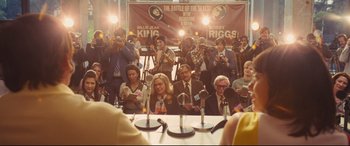 Movie still from “Battle of the Sexes” (2017), directed by Valerie Faris – A group of people sitting at a table with microphones in front of them; Wide shot, Over the shoulder angle