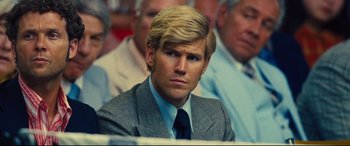 Movie still from “Battle of the Sexes” (2017), directed by Valerie Faris – A man in a suit and tie sitting in front of a group of people; Close Up shot, Over the shoulder angle